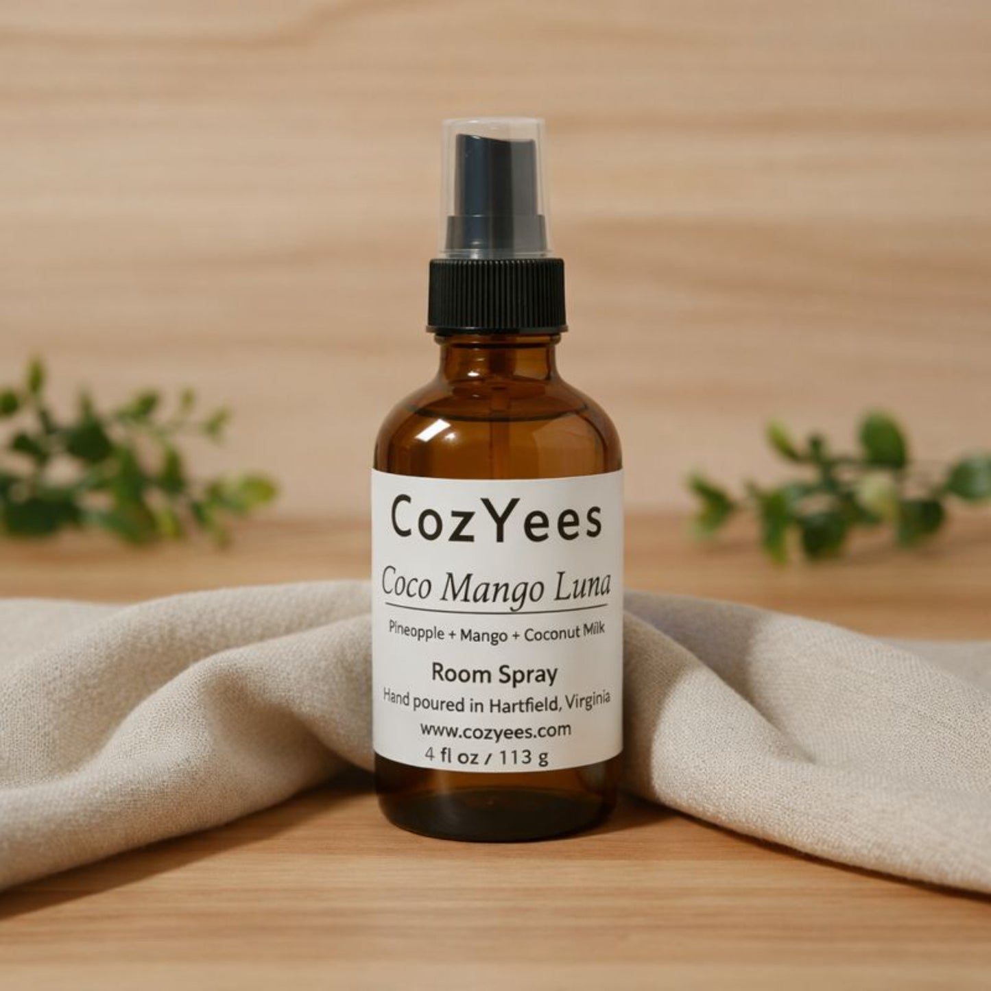 CozYees Coco Mango Luna room spray bottle on a wooden surface with a beige cloth and greenery in the background.