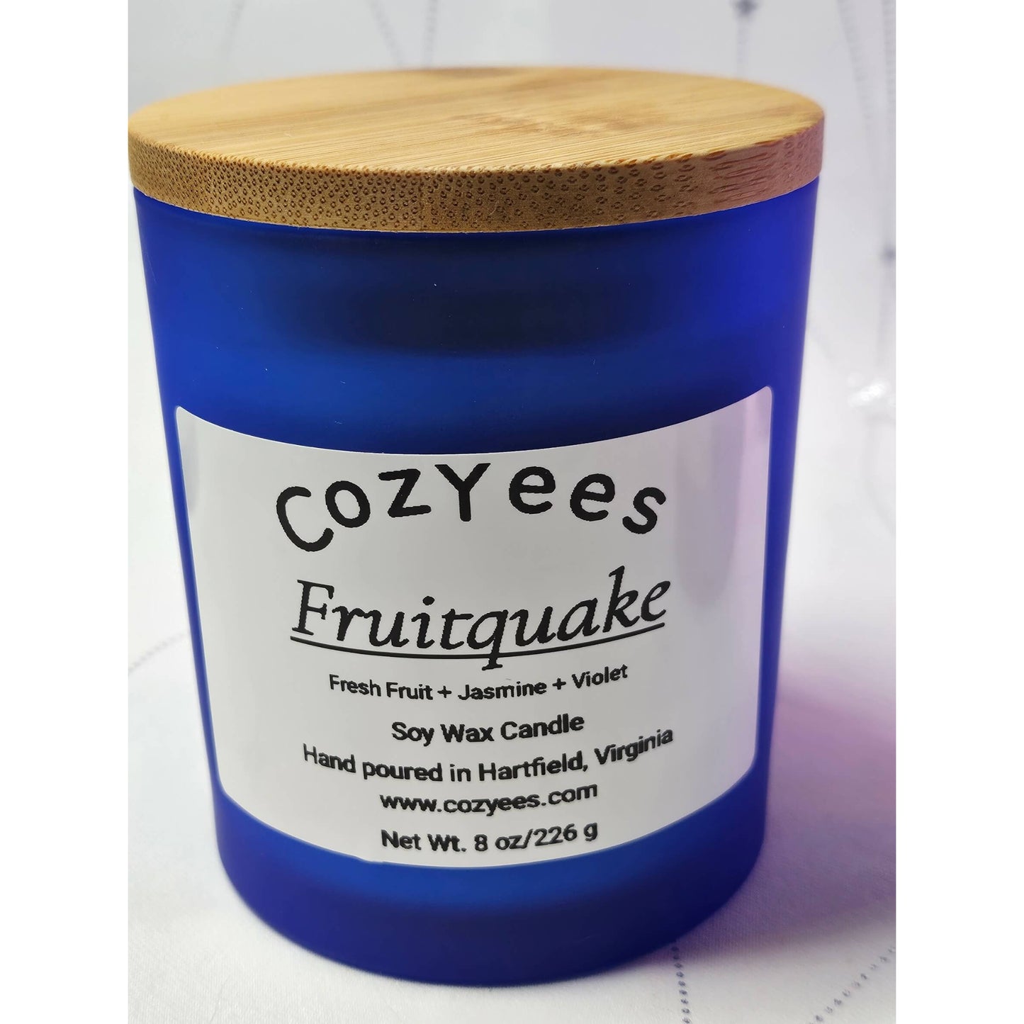 Blue candle with a wooden lid labeled 'Cozyees Fruitquake' on a white background