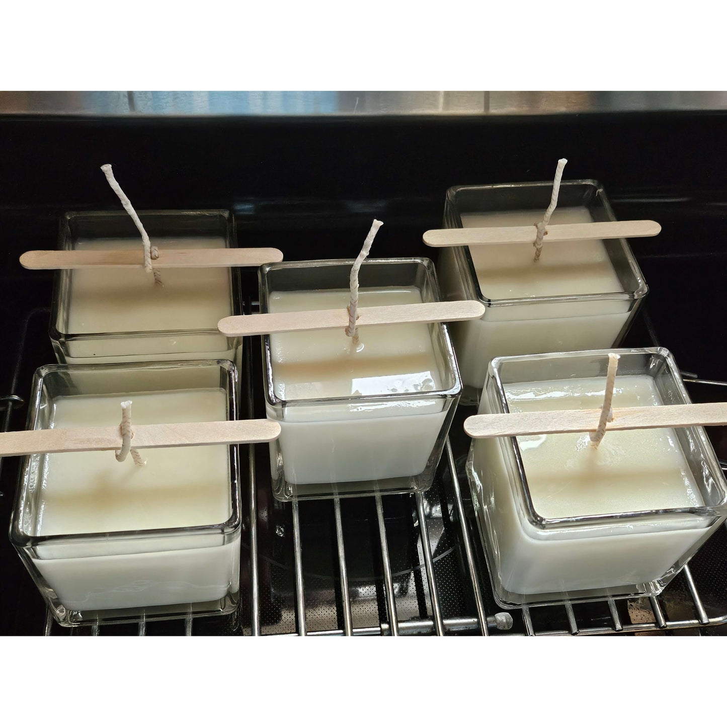 Multiple small square candles with wooden sticks on a metal rack.