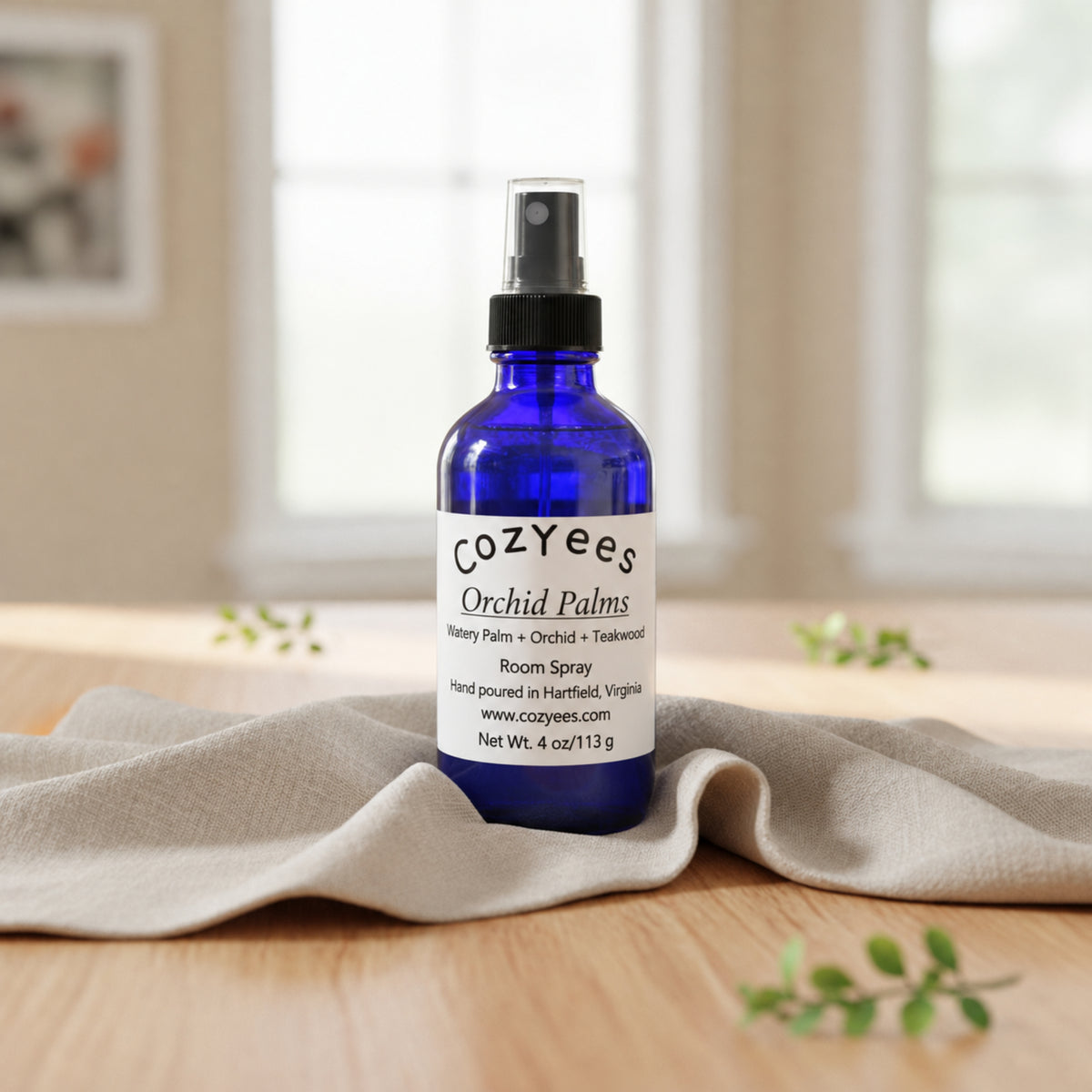 A cobalt blue glass room spray bottle with a black spray top and a white label reading 'Cozyees Orchid Palms', set on a beige cloth over a wooden surface with a softly blurred indoor background.