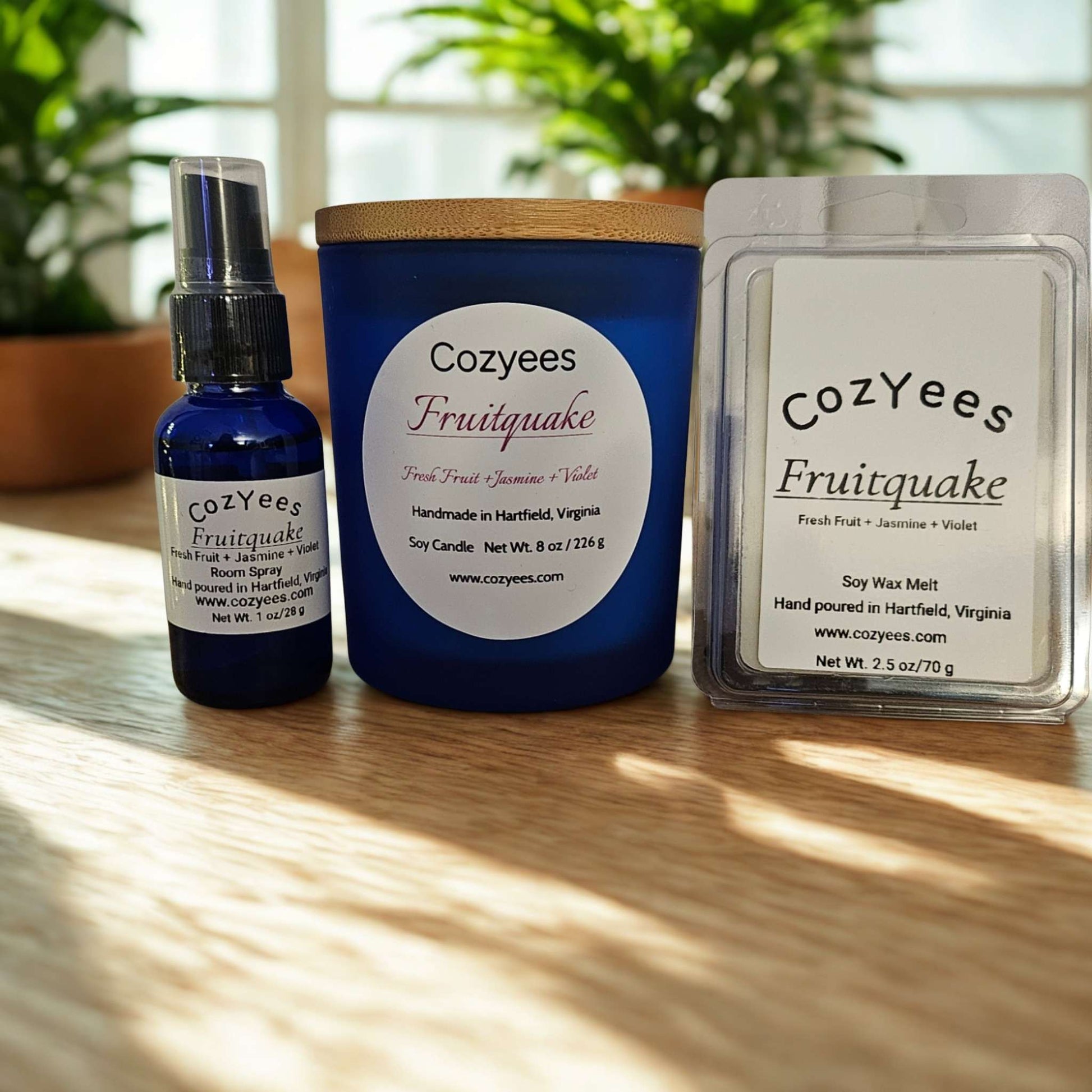 Cozyees Fruitquake candle, bottle, and wax melt on a wooden surface with plants in the background