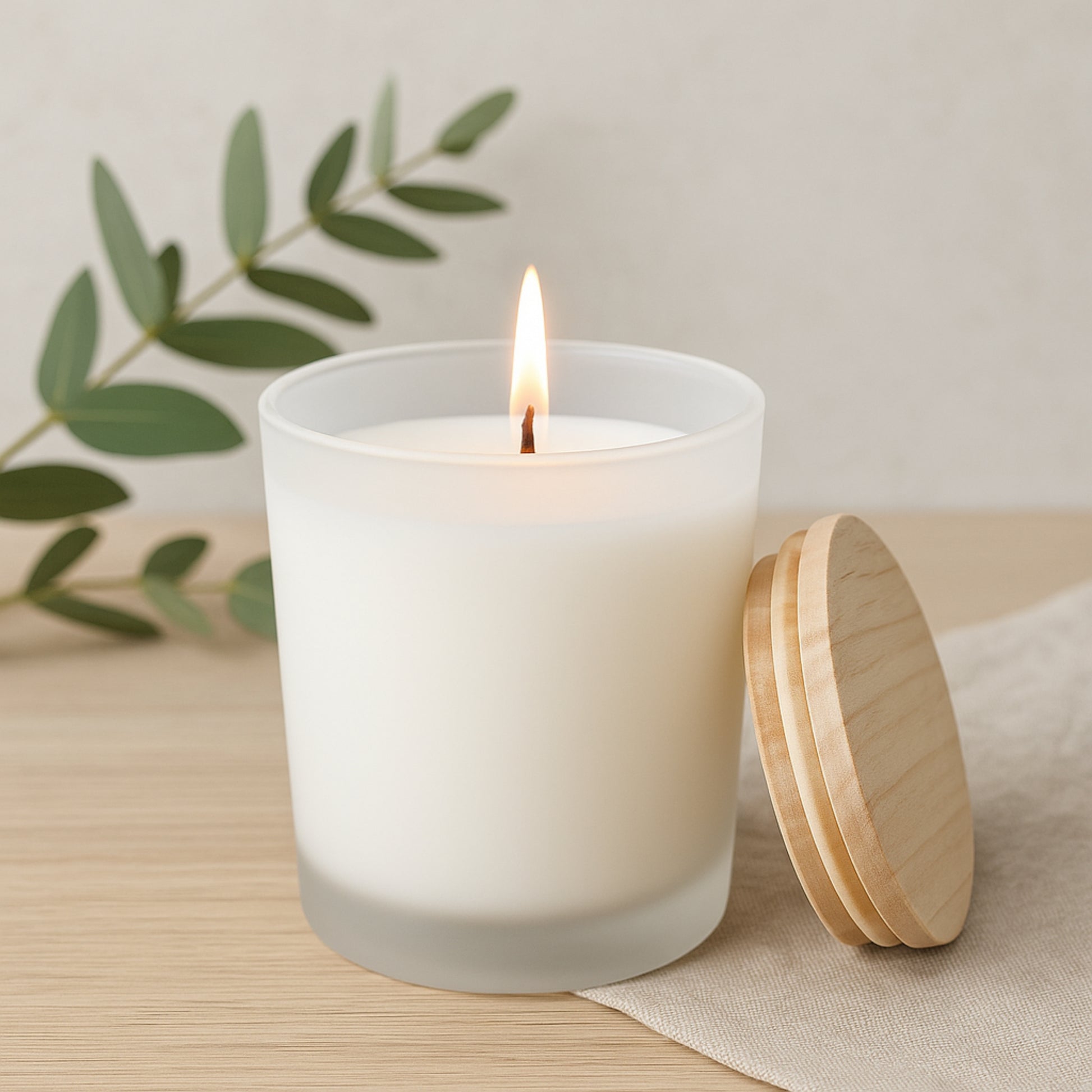 White candle with wooden lid on a light surface with green leaves in the background