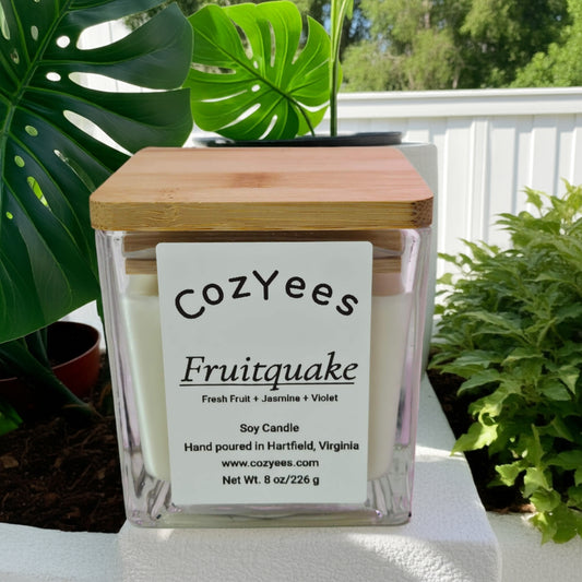 Cozyees Fruitquake candle with a wooden lid on a white surface with green plants in the background
