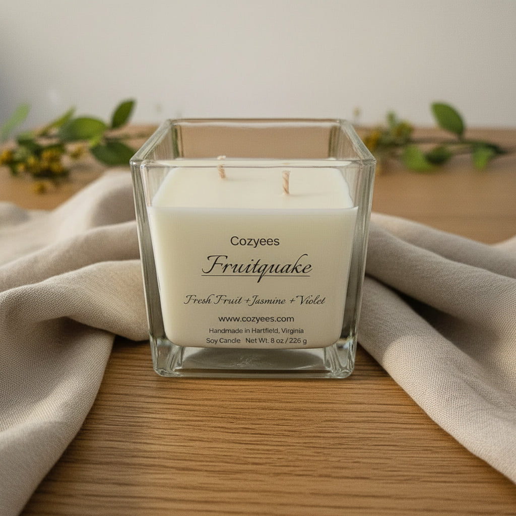 Cozyees Fruitquake candle in a clear square jar on a wood table.
