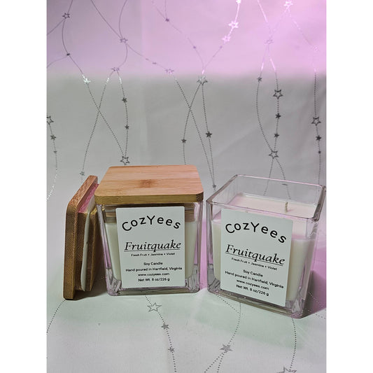 Two Cozyees Fruitquake candles, one in a wooden box and the other in a clear container, on a light gray background with star patterns.