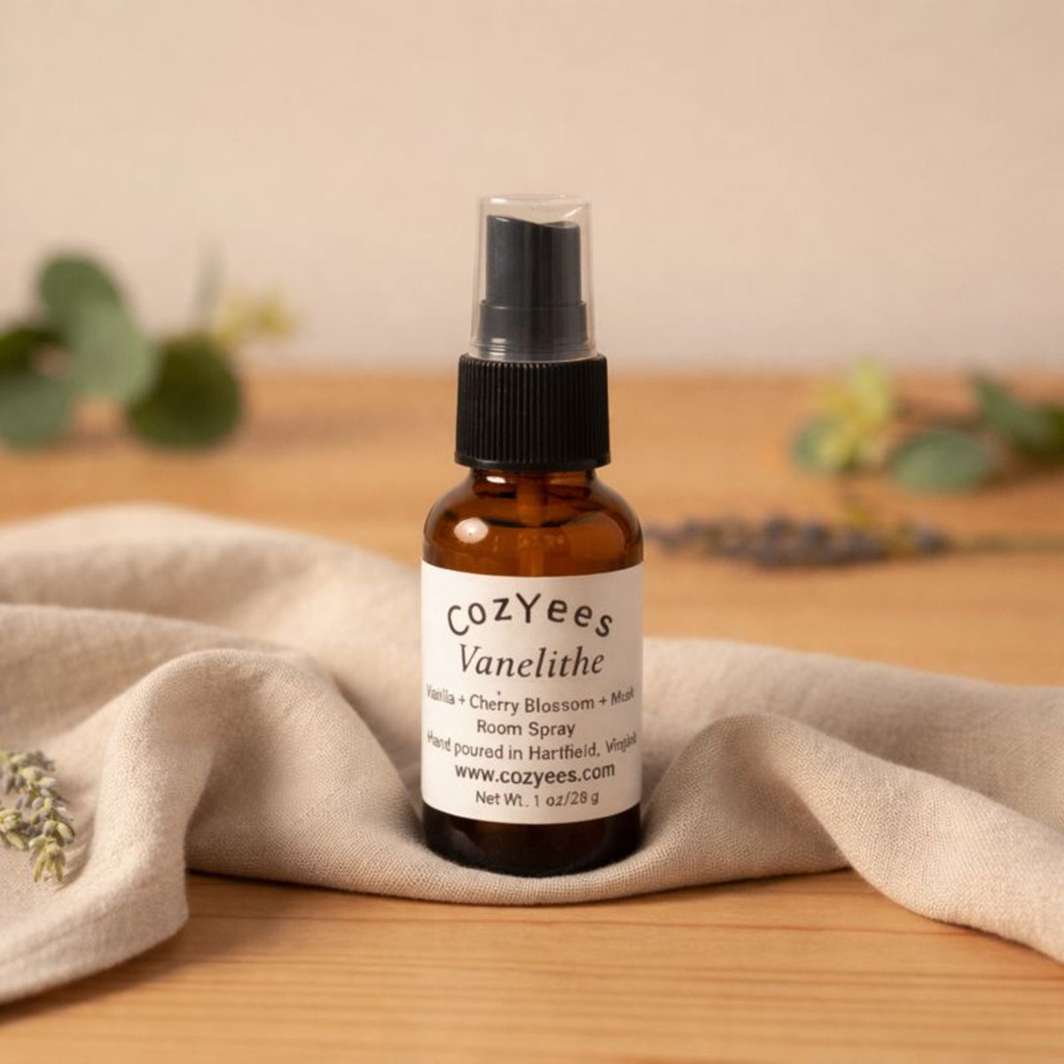 Cozyees Vanelithe room spray bottle on a wooden surface with a beige cloth and greenery in the background.