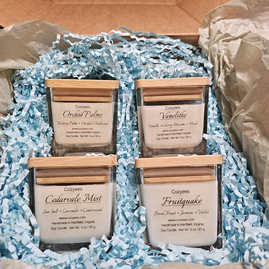 Cozyees Candle Sampler Gift Set: four 3 oz soy candles in square glass jars with bamboo lids, Orchid Palms, Vanelithe, Cedarvale Mist and Fruitquake, in a kraft box with blue crinkle paper and tissue