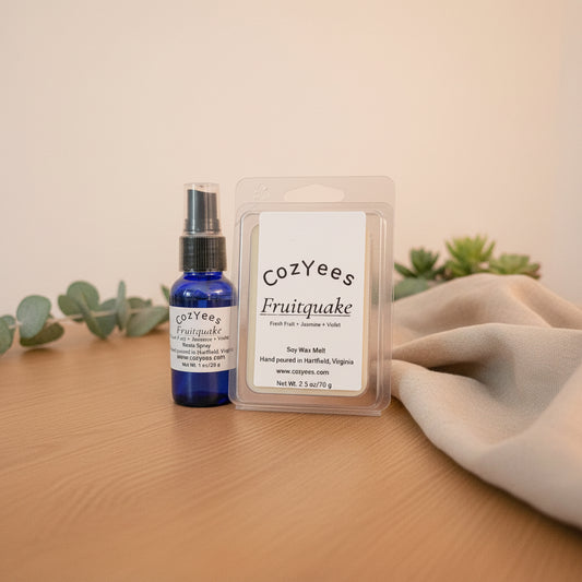 Cozyees Fruitquake room spray and soy wax melt set on a wooden surface in front of a dark background. The set includes a small cobalt blue room spray bottle and a clear plastic clamshell package containing a white wax melt card.