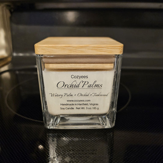 Close-up of a Cozyees Orchid Palms 3 oz soy candle in a square glass jar with a bamboo lid, scented with watery palm, orchid and teakwood