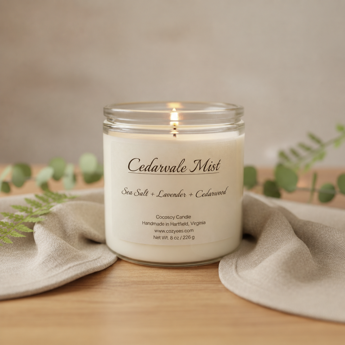 A lit jar candle labeled 'Cedarwale Mist' sits on a wooden surface draped with beige fabric, with soft green foliage in the background.