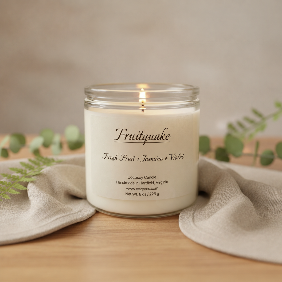 A lit glass jar candle labeled 'Fruitquake' sits on a wooden surface with draped linen cloth and soft greenery in the background. The candle is cream-colored wax in a clear glass container with a white label and visible flame.